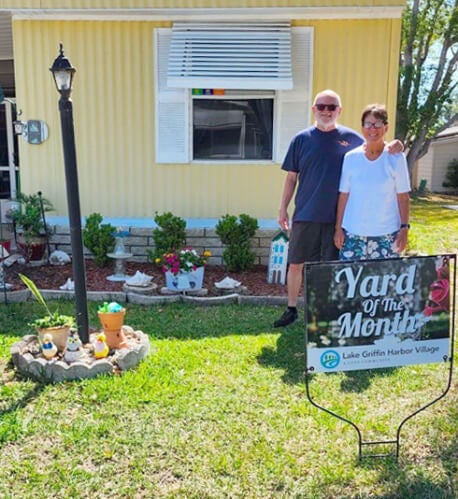 A 55+ couple standing in their front yard with a Yard of the Month sign at Lake Griffin Harbor Village in Leesburg, Florida.