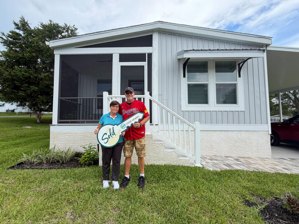 New 55+ homeowners on the front lawn at their new home with a large sign in the shape of a house key that says "SOLD!"