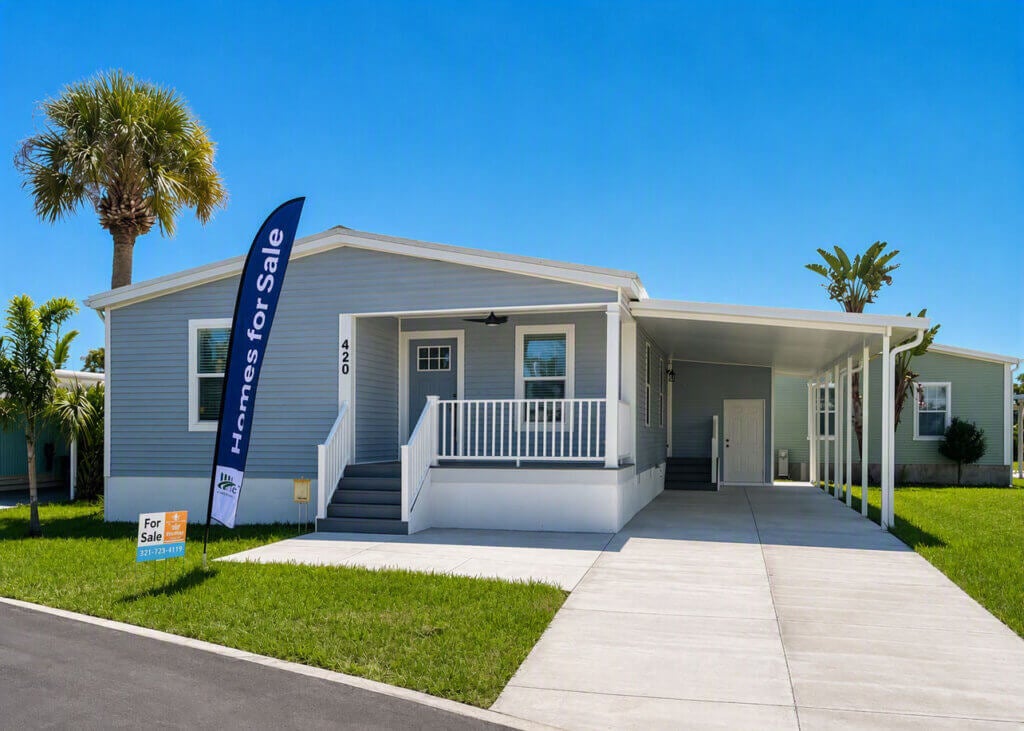 A manufactured home with a for sale sign in the front yard at The Waters in Melbourne Beach, Florida. The home features a front porch and car port.
