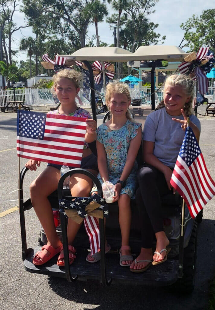 Three girls holding flags on the back of the golf cart during the Veterans Day golf cart parade.