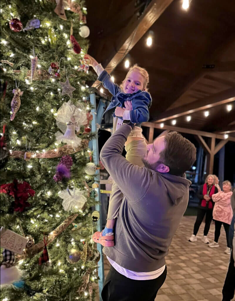 Father lifts up his daughter to place an ornament on the top of the Community Christmas tree