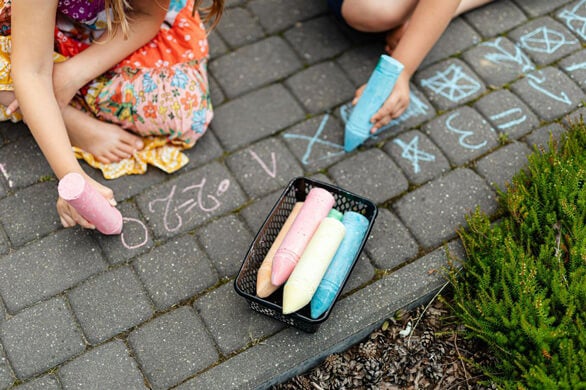 Two children drawing with chalk on a paved patio, with a basket of colorful chalk.