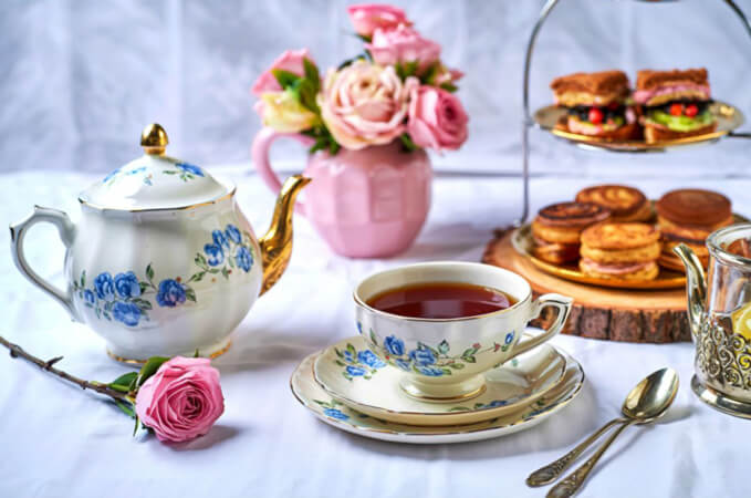 Floral teapot sitting next to a teacup and saucer, a pink rose, and a tiered stand with sandwiches and pastries.