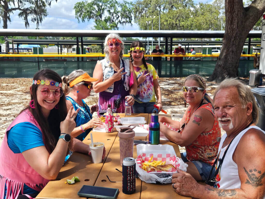 A group of people dressed in colorful tie-dye shirts and flower crowns sit at a picnic table, enjoying food and drinks at the Hippie Fest event at Holiday RV Park in Leesburg, FL.