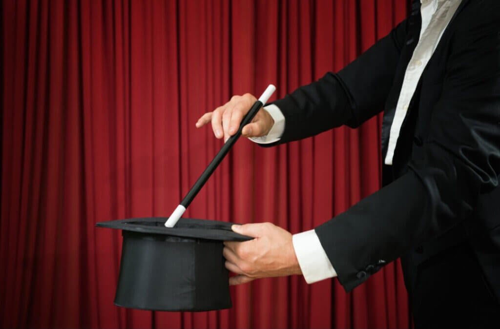 A person in a suit holds a wand over a black top hat, against a red curtain backdrop.