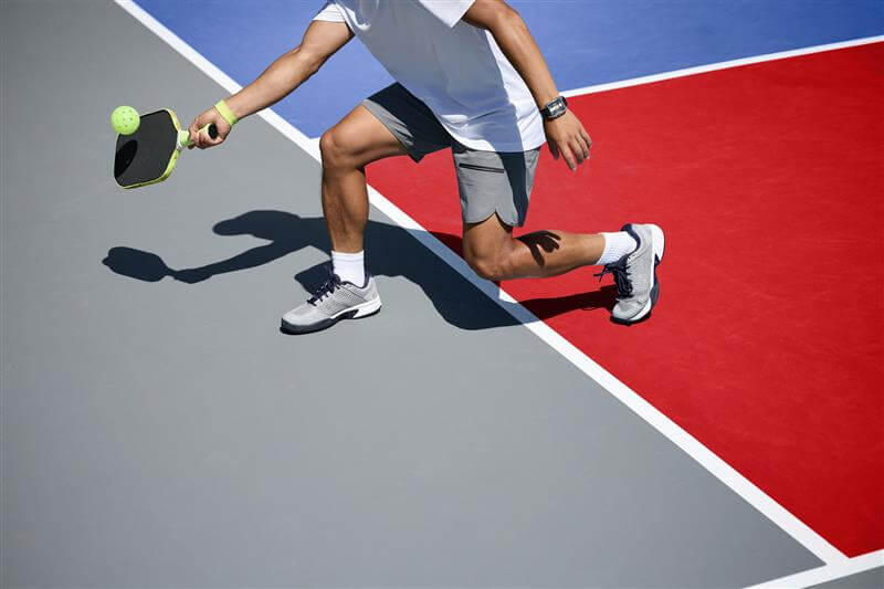 Generic pickball court image of a man hitting a pickleball on a gray and red court.