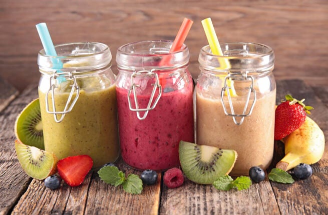 Three glass jars filled with colorful smoothies, garnished with fresh fruits and mint leaves on a wooden surface.
