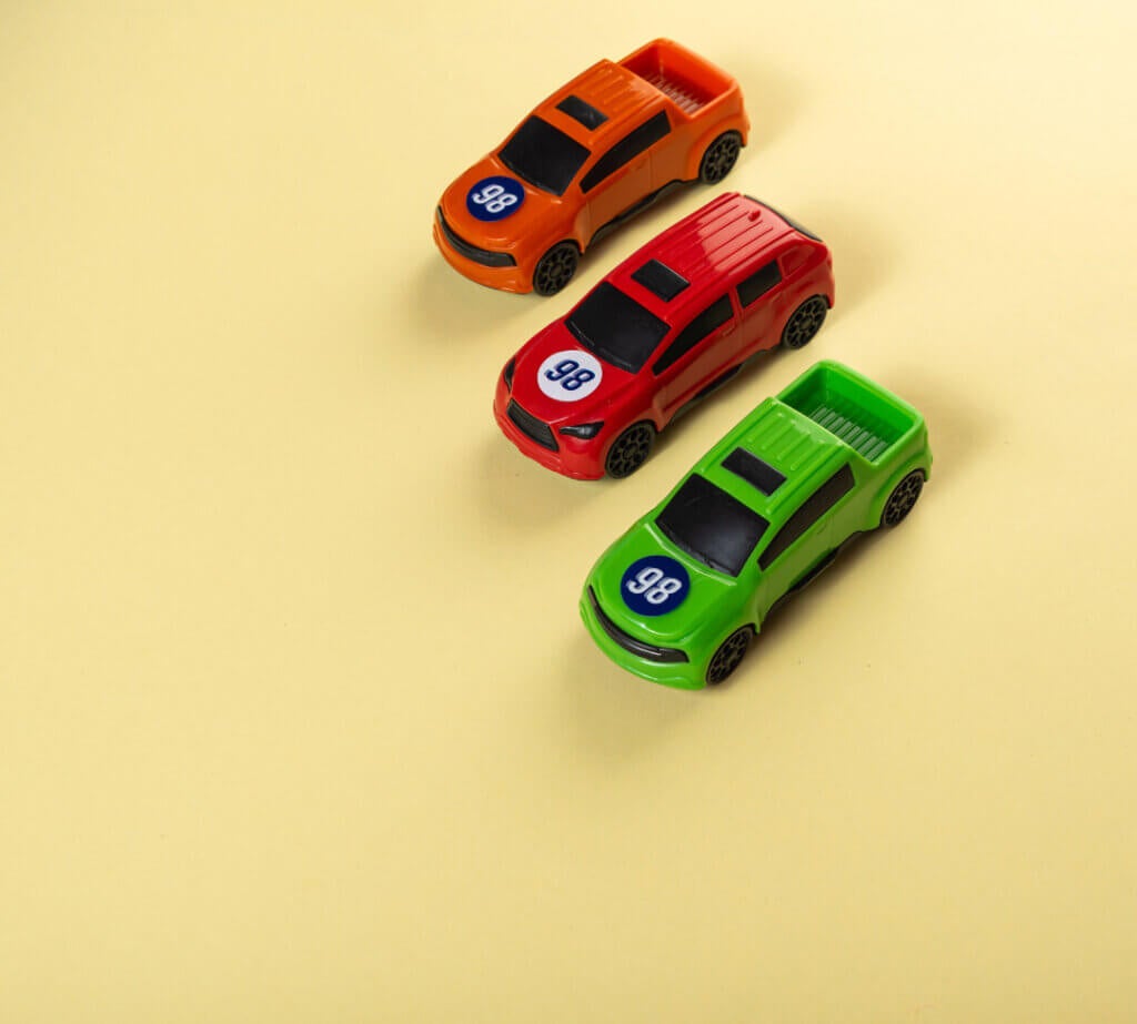 Three toy cars, orange, red, and green, are lined up diagonally on a pale yellow background.