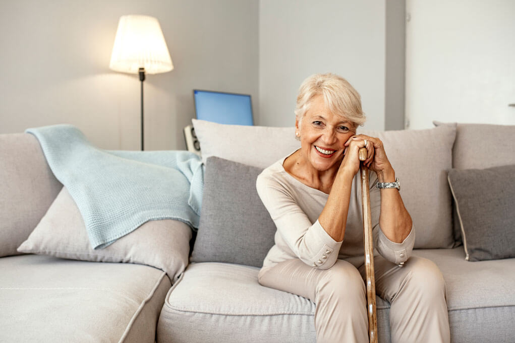 Retired short hair old woman with her walking stick at home looking at camera during the day while sitting on sofa. Senior gray hair women supporting on a walking cane at home.