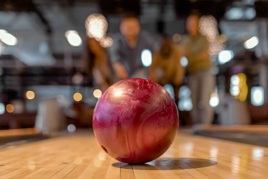 Detailed foreground focus on bowling ball on bowling alley lane, with friends having playful bowling session blurred in background. Represents fun, friendship, competition and entertainment venue atmosphere.
