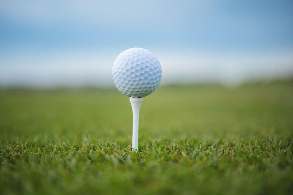 A close-up shot of a golf ball positioned on a tee, surrounded by lush green grass and under a gentle blue sky. The image captures a serene and focused moment in the game of golf, emphasizing tranquility and precision. Credit: iStock
