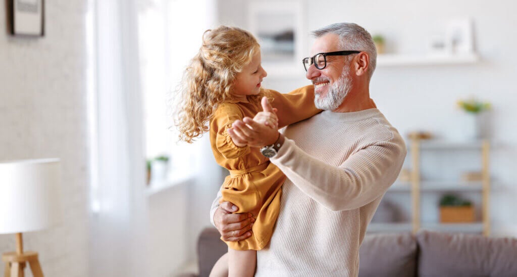 Elegant loving caring grandfather looking at his cute little granddaughter, adorable child girl and positive grandpa holding hands while dancing together in living room at home. Family concept