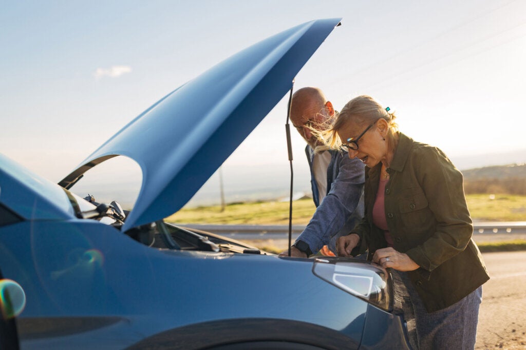 Senior couple beside a stopped car with open hood, examining the engine and trying to solve the issue