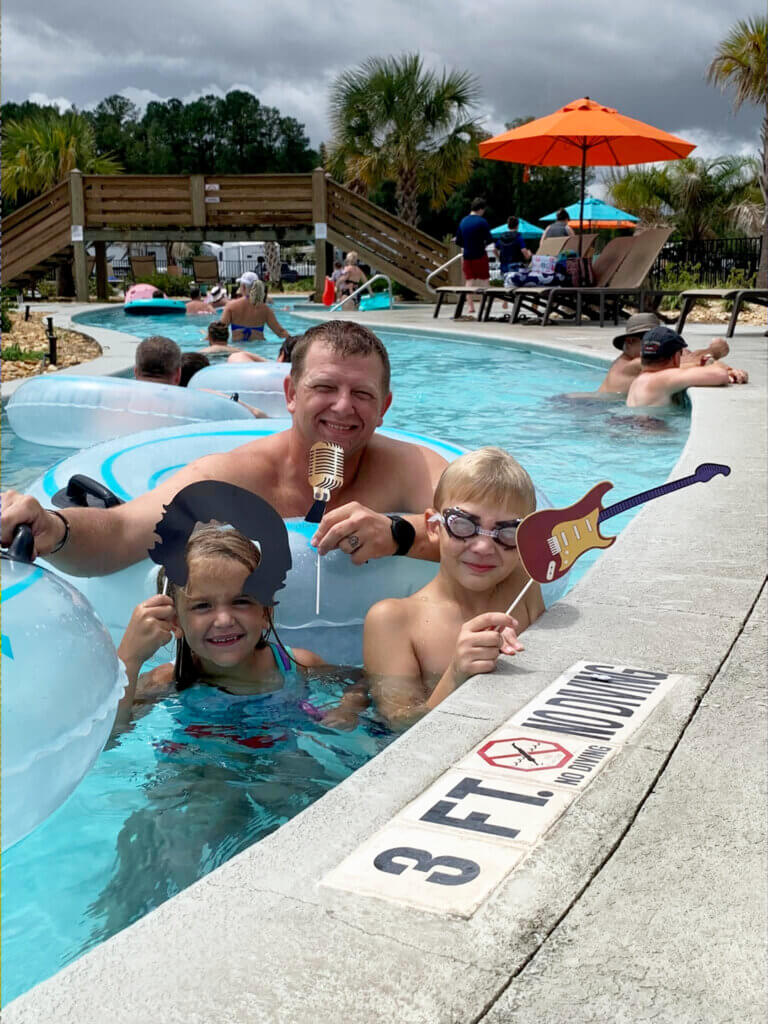 Father and 2 kids hold guitar, microphone, and rocker hair photo props while in the lazy river at CreekFire RV Park.