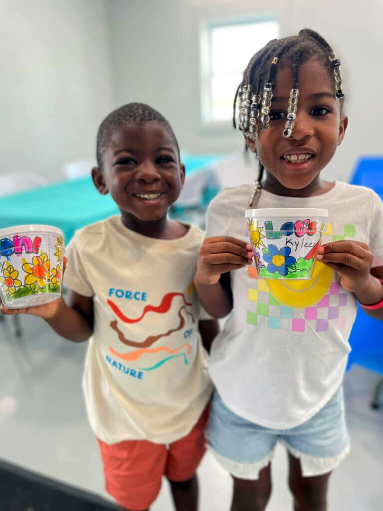 Two children at CreekFire RV proudly show decorated cups with colorful drawings of flowers and names from planned activity.