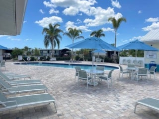 View of the outside swimming pool at Estero Bay Village in Florida. The pool deck is complete with r tables with ubrellas and chairs and many Lounge reclining chairs linin gthe perimeter of the pool enclosure.
