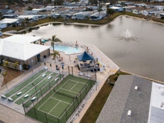 Drone view of the lake within The Meadows surrounded by manufactures homes, the clubhouse, swimming pool, shuffleboard courts, and pickleball courts.