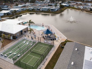 Drone view of the lake within The Meadows surrounded by manufactures homes, the clubhouse, swimming pool, shuffleboard courts, and pickleball courts.