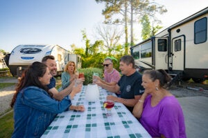 Six friends cheersing with drinks at a picnic table between two RVs at CreekFire RV Resort in Georgia.