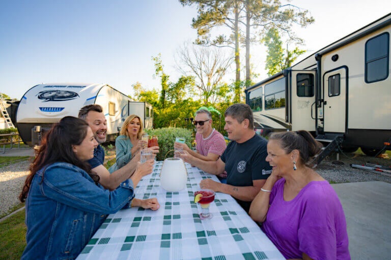 Six friends cheersing with drinks at a picnic table between two RVs at CreekFire RV Resort in Georgia.