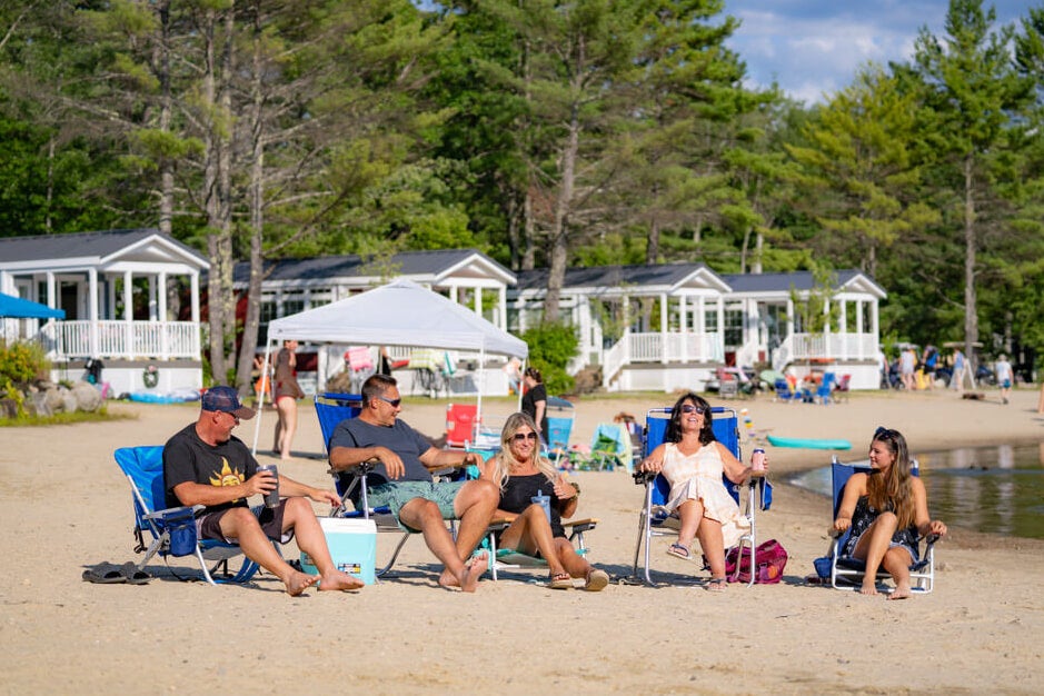 Friends sitting on the beach at Point Sebago Resort having drinks and chatting with cabins in the background.