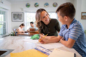 Mom doing homework with her son at the kitchen counter. A father and daughter at the kitchen table in the background.