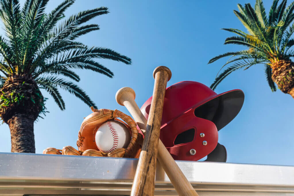 Looking up at a new baseball in a brown leather glove along with a red batting helmet and two wooden baseball bat on an aluminum bench with a palm tree and blue sky in the background for Spring Training. Photo from iStock.