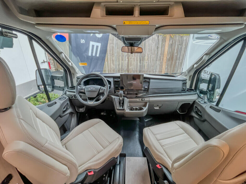 Leisure Travel Van Wonder front Interior view of a Leisure Travel camper van showing two light-colored leather front seats, a dashboard with a large touchscreen, steering wheel, and gear shifter.