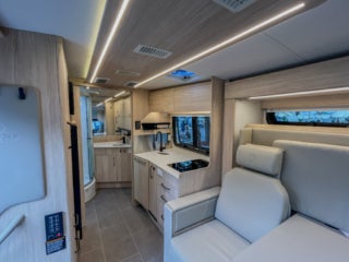 Interior view of a Wonder Camper Van kitchen with light wood cabinets, a sink, stovetop, and a window. A light-colored sofa is visible to the right.