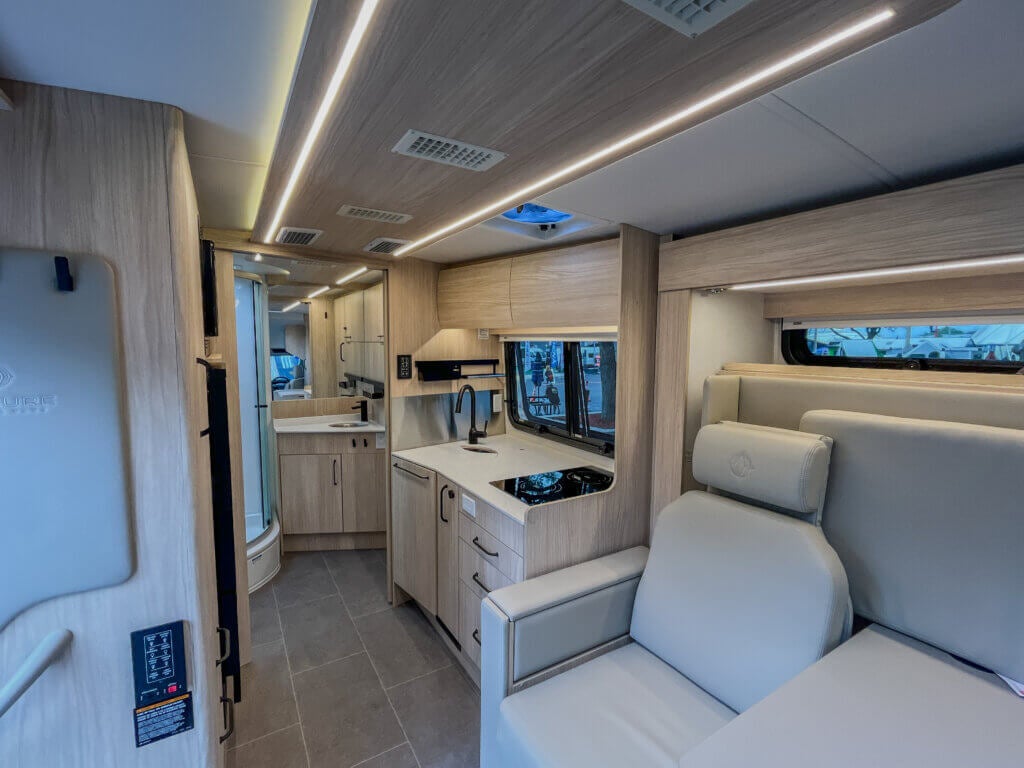 Leisure Travel Van Wonder Interior view of a Wonder Camper Van kitchen with light wood cabinets, a sink, stovetop, and a window. A light-colored sofa is visible to the right.