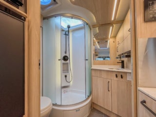A Leisure Travel Camper Van bathroom with a curved glass shower stall, a toilet, and a vanity with wood-grain cabinets and a white countertop.