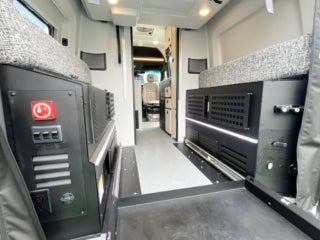 Interior view of a 2026 GH1 Tiffin Class B model Camper van conversion showing two seats with patterned upholstery, black cabinets with vents and switches, and a walkway leading to the driver's area.