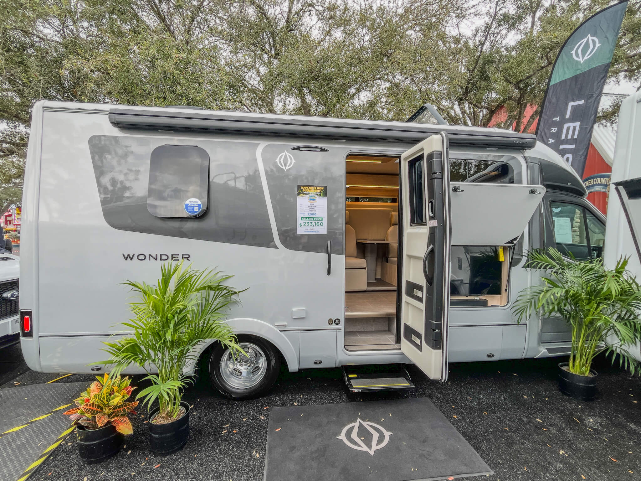Leisure Travel Van Wonder Gray 02 Gray Leisure Travel Camper Van parked outdoors with its doors open, revealing an interior with seating and a table. Potted plants are placed around the vehicle.