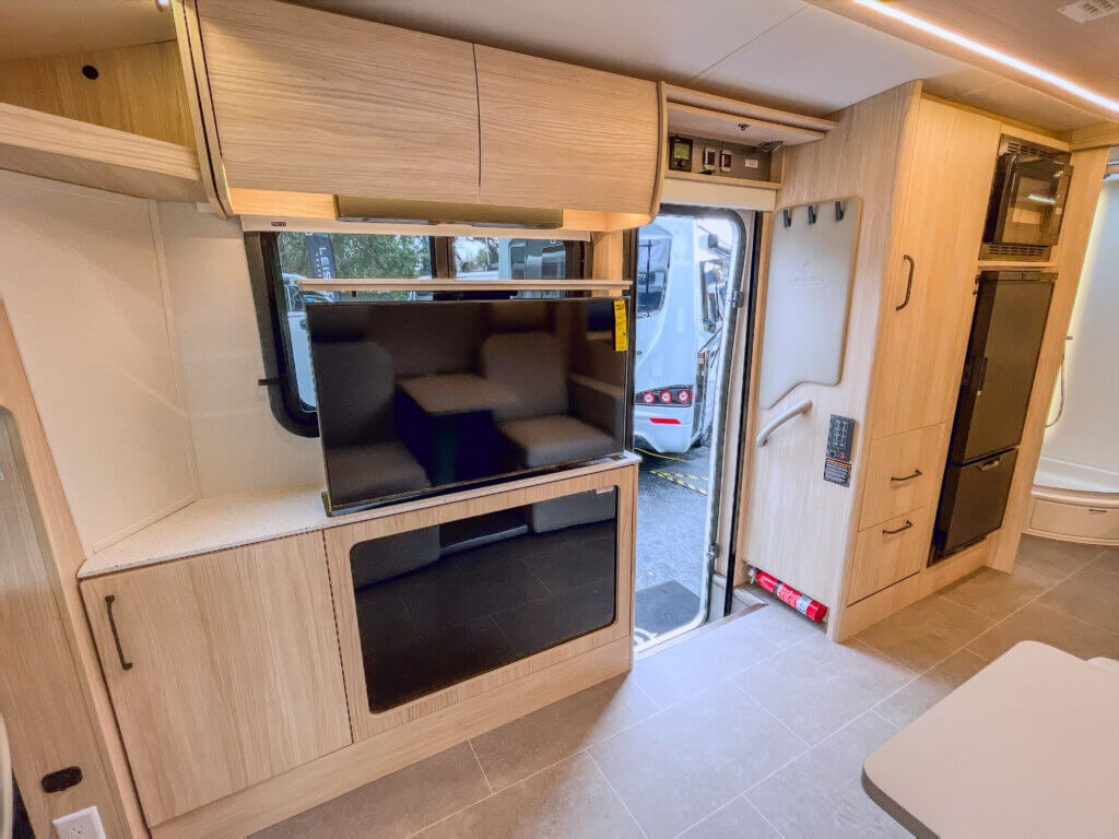 Leisure Travel Wonder Camper Interior view of a Leisure Travel camper van, showing a mounted television, kitchen cabinets, a refrigerator, and a microwave above the refrigerator.