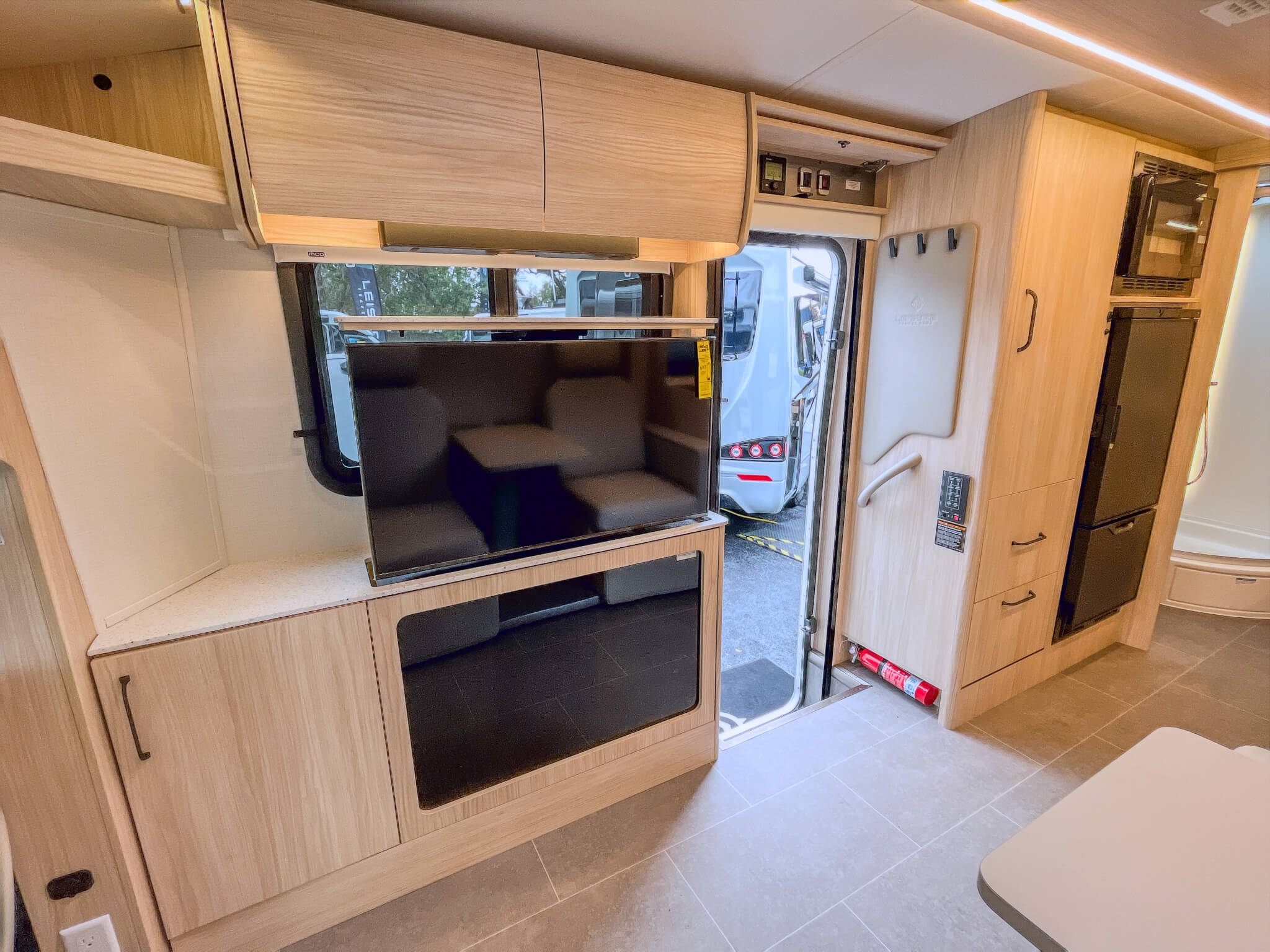 Leisure Travel Wonder Camper Interior view of a Leisure Travel camper van, showing a mounted television, kitchen cabinets, a refrigerator, and a microwave above the refrigerator.