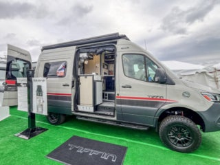 A Tiffin camper van with its door open, revealing the interior, parked on artificial grass with a Tiffin logo mat in front.