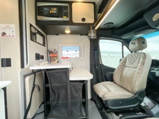 Interior view of a Tiffin camper van, showing a compact area with a microwave, overhead cabinets, and audio controls leading to the driver's cabin.