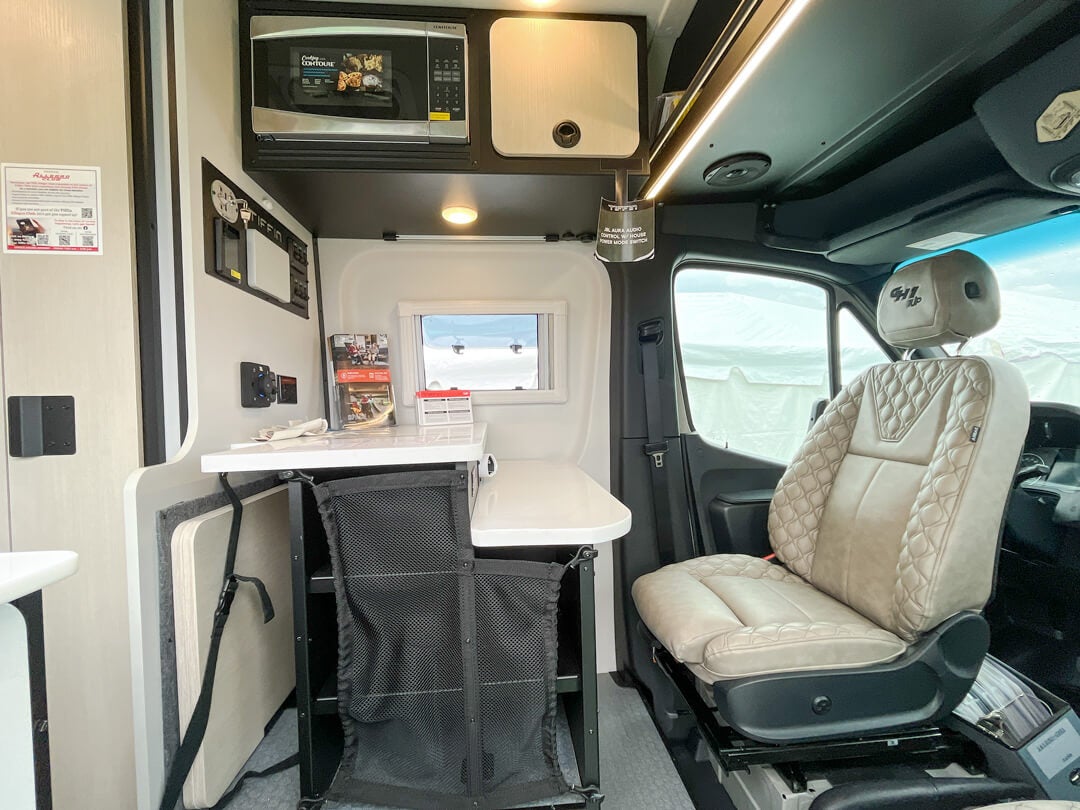 Tiffin Camper Van GH1 09 Interior view of a Tiffin camper van, showing a compact area with a microwave, overhead cabinets, and audio controls leading to the driver's cabin.