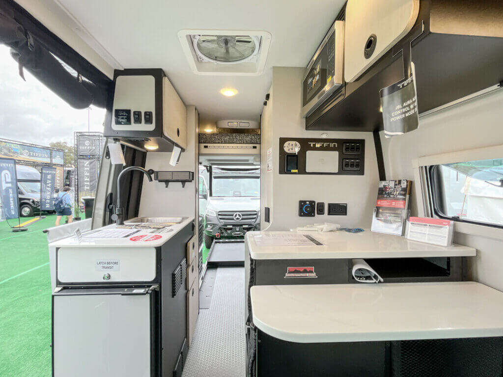 Tiffin Camper Van GH1 12 Interior view of a Tiffin camper van, showing a compact kitchen area with a sink, refrigerator, and overhead cabinets, leading to the driver's cabin.