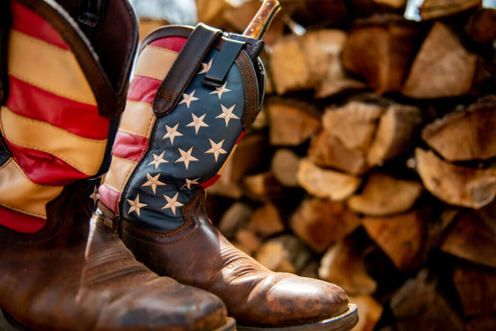 Western boots with an American flag pattern in front of a pile of chopped wood.