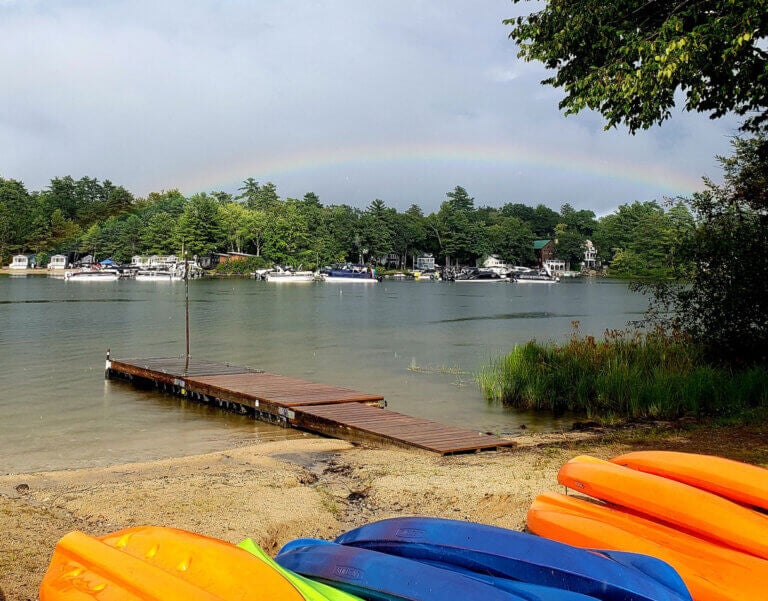 Colorful kayaks along the shore of Sebago Lake in Maine with Point Sebago Resort cabins and a rainbow in the backrgound.