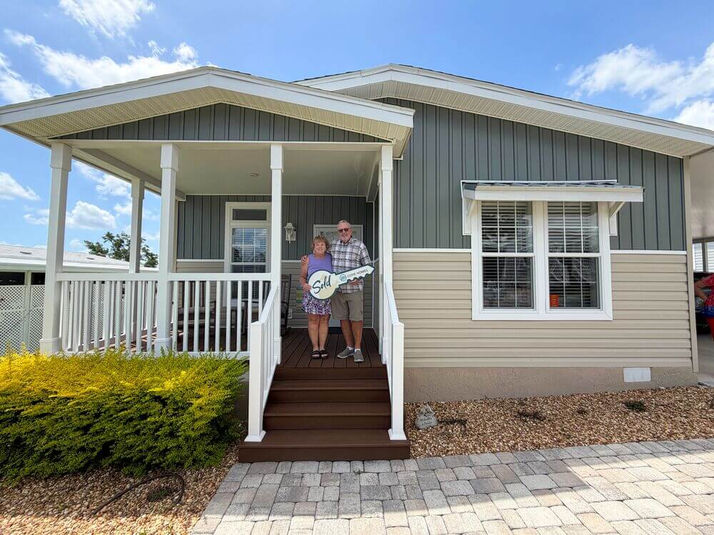 RGV New Homeowners 3 New 55+ homeowners on the front porch of their new home with a large sign in the shape of a house key that says "SOLD!"
