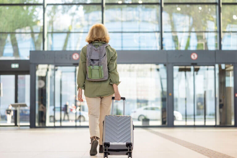 An older woman with a backpack and pulling a suitcase is walking into the airport.