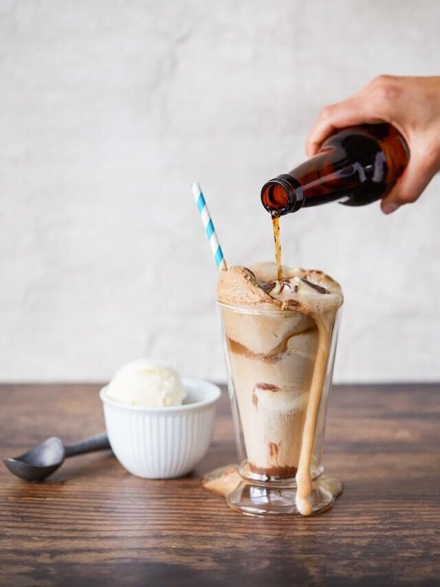 Root beer being poured into a rootbeer float with a scoop of vanilla ice cream in the background.
