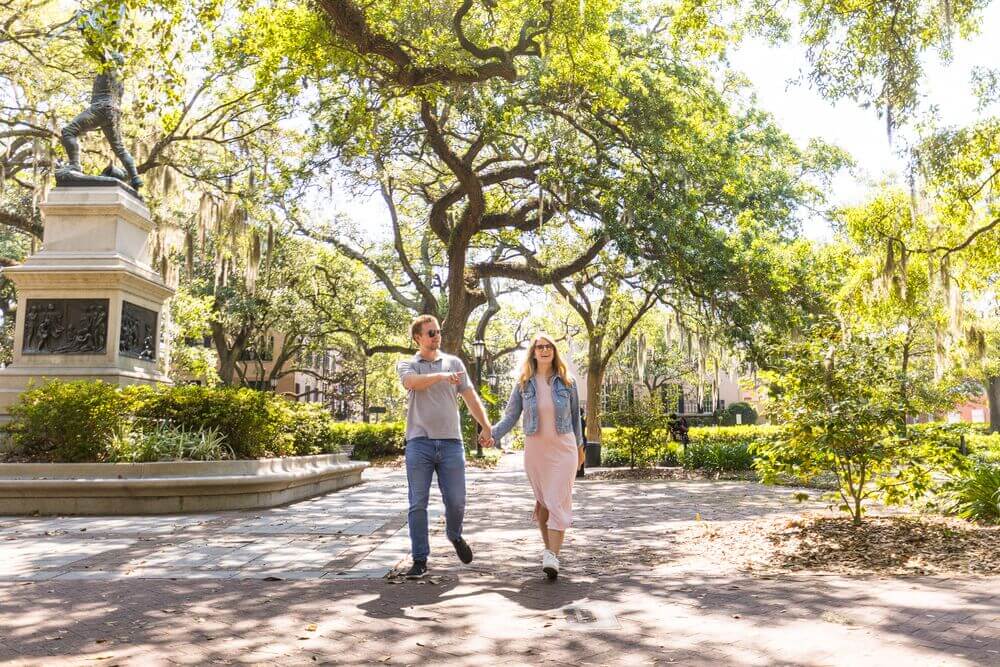 A couple walks through an historic Savannah, Georgia park with a statue on the left.