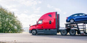Industrial car hauler red big rig semi truck tractor with extended cab for truck driver rest transporting cars on modular semi trailer driving on highway road at sunny day