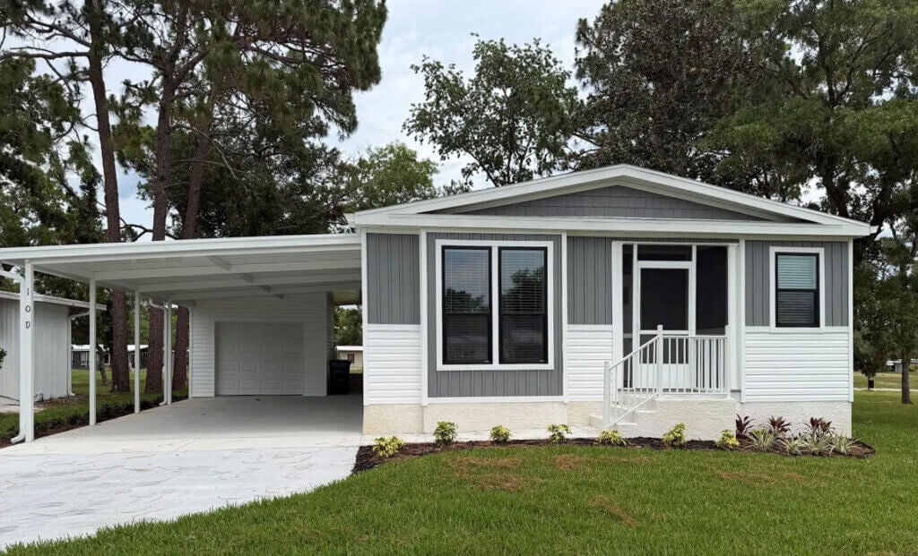A gray and white manufactured home for sale in Ocala, Florida at Rolling Greens Village. The home features a spacious lawn and a carport.