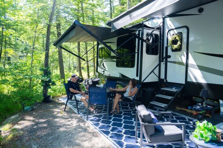 A fifth wheel trailer parked at a seasonal campsite on Sebago Lake in Maine with two people sitting outside.