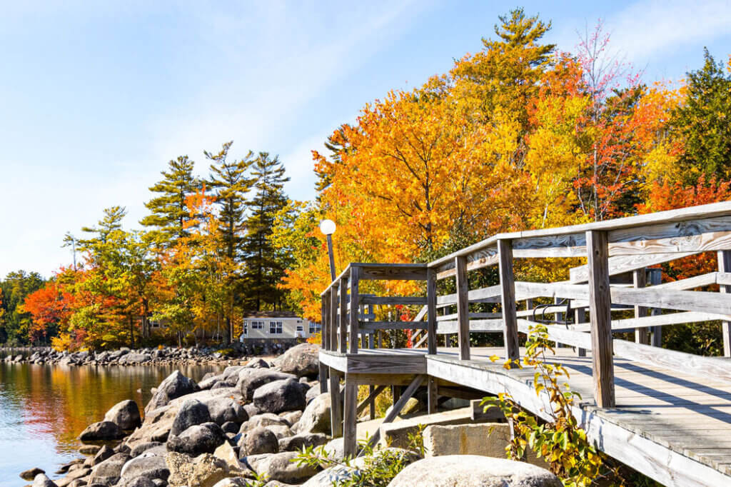 The fall foliage at Point Sebago Resort in Casco, Maine, with a wooden walkway over rocks leading to cabins on Sebago Lake.
