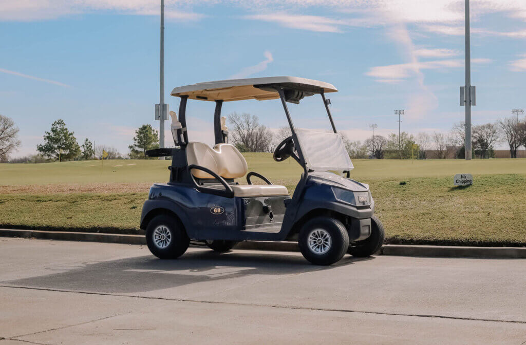 A golf cart parked on a pathway next to a golf course under a clear blue sky.